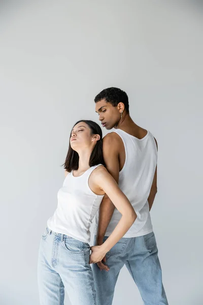 Чувственная азиатка с закрытыми глазами, опирающаяся на африканского американца в белой майке и джинсах, изолированных на сером — стоковое фото