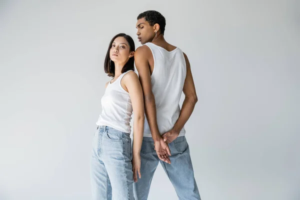 Asian woman in white tank top and blue jeans looking at camera near young african american guy isolated on grey — Stock Photo