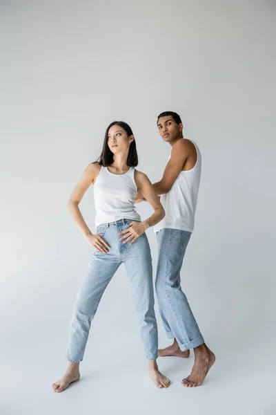 Full length of stylish asian woman posing with thumbs in pockets of jeans near barefoot african american man on grey background — Stock Photo