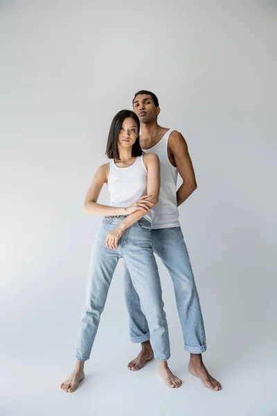 Longitud completa de la pareja multiétnica descalza y delgada en camisetas sin mangas y pantalones vaqueros sobre fondo gris - foto de stock