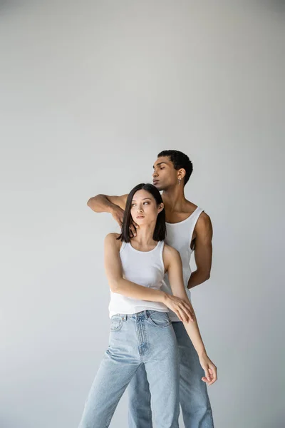 Junger afrikanisch-amerikanischer Kerl berührt Hals einer asiatischen Frau in weißem Tank-Top und Jeans isoliert auf grau — Stockfoto