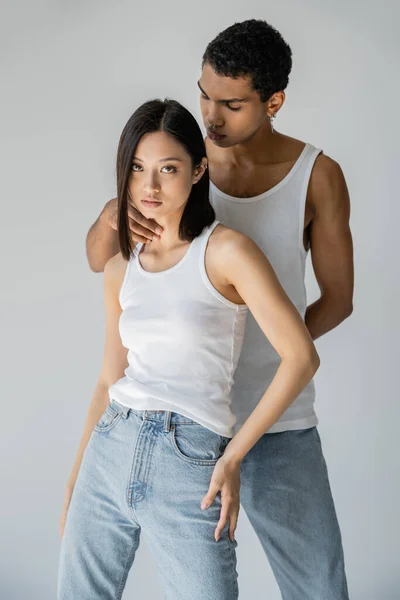 Joven africano americano hombre tocando cuello de asiático mujer en blanco tanque top y jeans aislado en gris - foto de stock