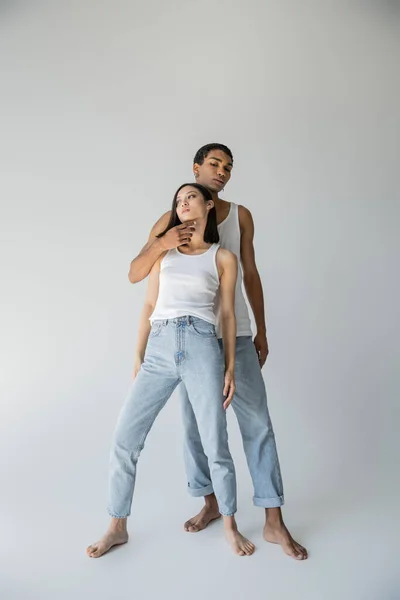 Full length of stylish african american man touching chin of barefoot asian woman in tank top and jeans on grey background — Stock Photo