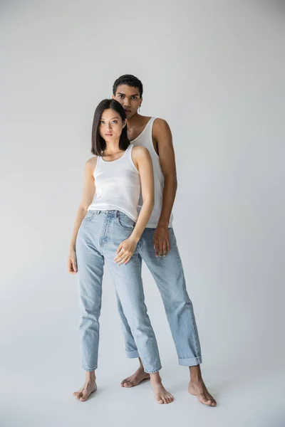 Полная длина молодых и босых межрасовых пар, позирующих в белых топах и синих джинсах на сером фоне — стоковое фото
