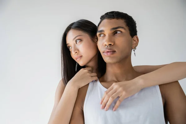 Sensual asian woman embracing stylish african american man in white tank top while looking away isolated on grey — Stock Photo