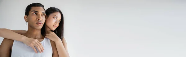 Brunette asiatique femme câlin élégant afro-américain gars en blanc débardeur isolé sur gris, bannière — Stock Photo