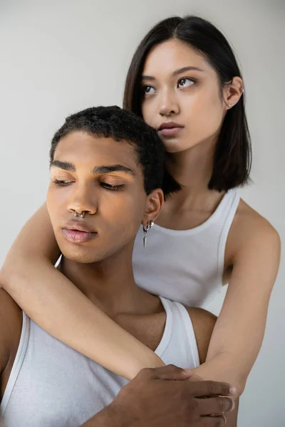 Charming asian woman hugging young african american man with silver piercing isolated on grey — Stock Photo
