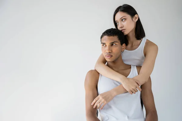 Young asian woman looking away and hugging african american man in white tank top isolated on grey — Stock Photo