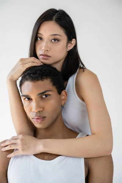 Sensual asian woman in white tank top embracing african american man with silver piercing isolated on grey — Stock Photo