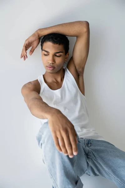 Jeune homme afro-américain en débardeur blanc et jeans assis sur fond gris — Photo de stock