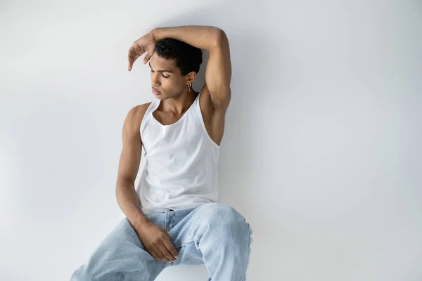 Задумчивый африканский американец в белом топе и синих джинсах, сидящих на сером фоне — стоковое фото