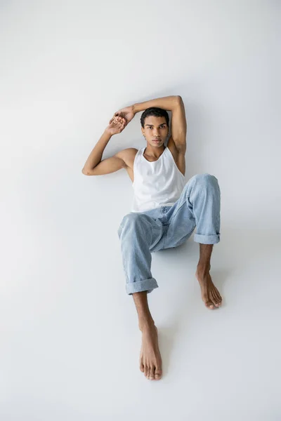 Tutta la lunghezza del ragazzo afroamericano a piedi nudi in jeans blu e canotta seduta e guardando la fotocamera su sfondo grigio — Foto stock