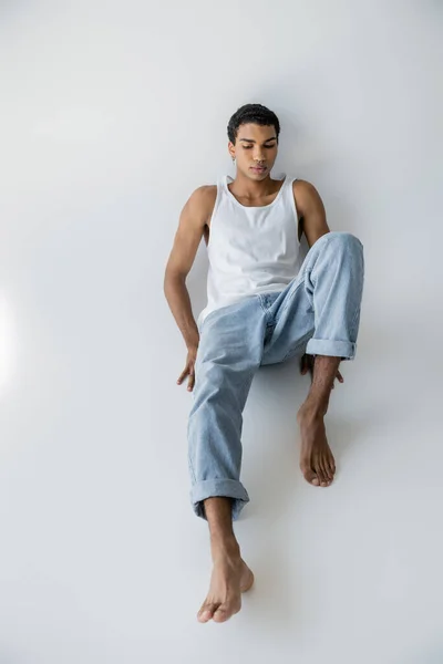 Vista de ángulo alto de hombre afroamericano joven y descalzo posando en camiseta sin mangas y jeans sobre fondo gris - foto de stock