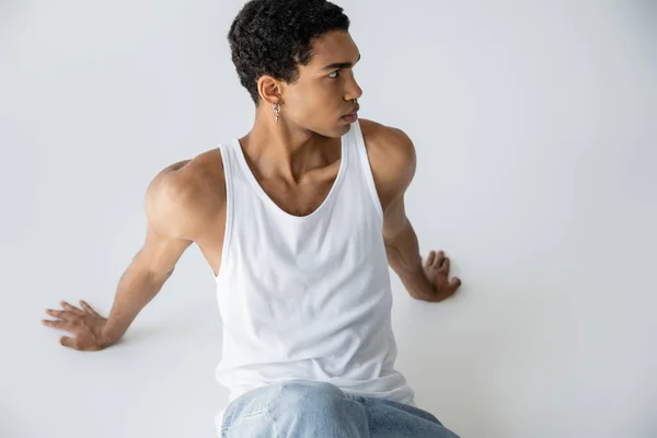 Afrikanisch-amerikanischer Mann in Tank Top und Blue Jeans schaut weg, während er auf grauem Hintergrund sitzt — Stockfoto