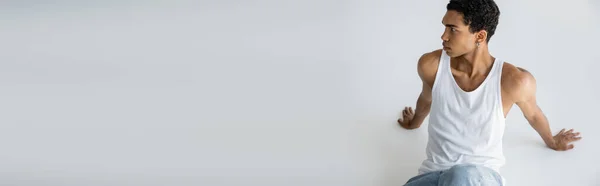 Élégant homme afro-américain en débardeur blanc assis et regardant loin sur fond gris, bannière — Photo de stock