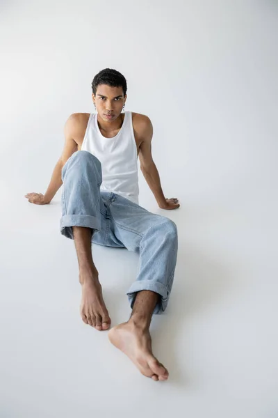 Piena lunghezza di elegante uomo africano americano in jeans blu e canotta seduta e guardando la fotocamera su sfondo grigio — Foto stock