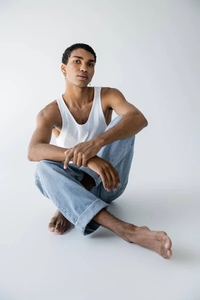 Descalzo afroamericano hombre en pantalones vaqueros azules sentado con las piernas cruzadas y mirando a la cámara sobre fondo gris - foto de stock