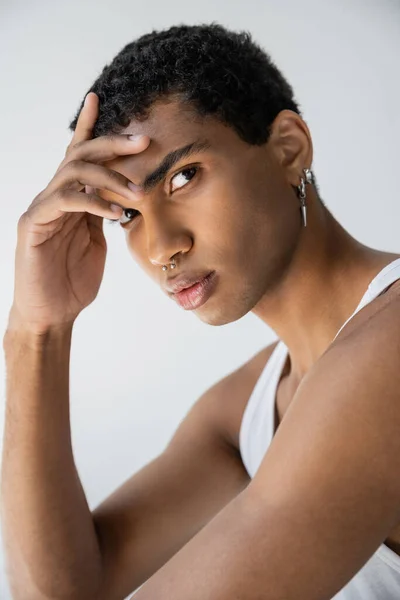 Retrato de hombre afroamericano con piercing plateado sosteniendo la mano cerca de la frente y mirando a la cámara aislada en gris - foto de stock