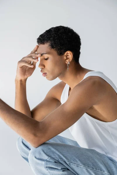 Side view of young african american man in white tank top and jeans touching forehead while sitting on haunches isolated on grey — Stock Photo