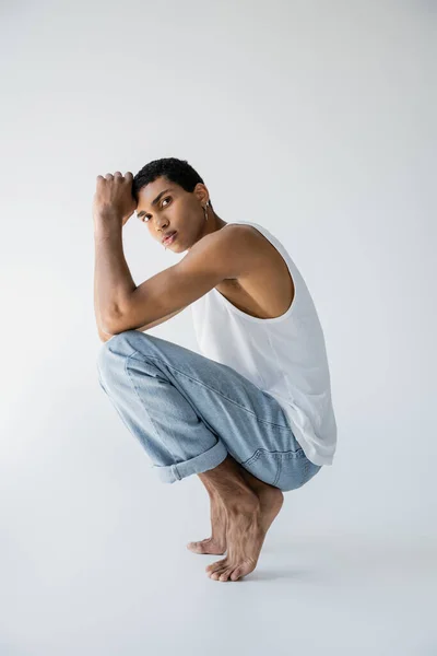 Barefoot and trendy african american guy posing on haunches and looking at camera on grey background — Stock Photo