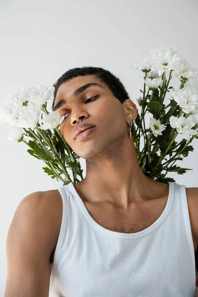 Retrato de un joven afroamericano en camiseta blanca posando con crisantemos blancos aislados en gris - foto de stock