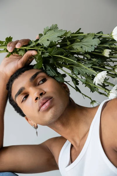 Young african american man in white tank top posing with white chrysanthemums isolated on grey — Stock Photo