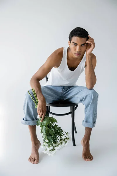 Volle Länge der barfuß und stilvolle afrikanisch-amerikanische Mann mit weißen Blumen sitzt auf Stuhl und blickt in die Kamera auf grauem Hintergrund — Stockfoto