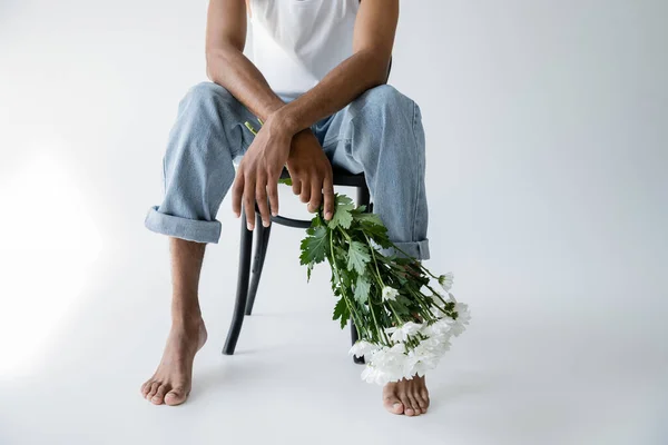 Обрізаний вид босоніж афроамериканця в джинсах, що тримає білі квіти, сидячи на стільці на сірому фоні — стокове фото
