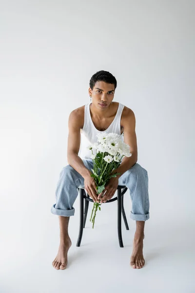Tutta la lunghezza dell'uomo afroamericano in canotta e jeans che reggono i fiori e guardano la macchina fotografica mentre si siede su sfondo grigio — Foto stock