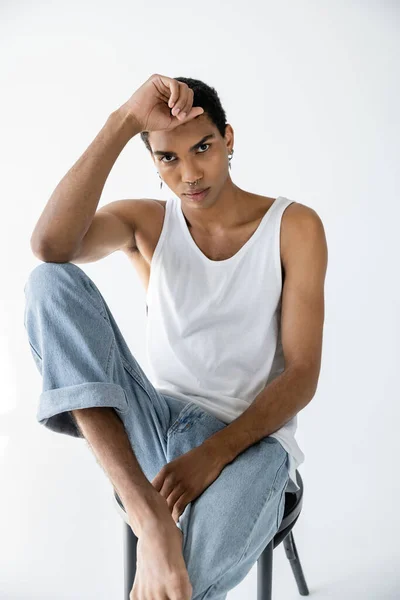 Trendiger afrikanisch-amerikanischer Mann in Jeans und Tank-Top posiert auf Stuhl und blickt in die Kamera auf grauem Hintergrund — Stockfoto