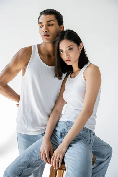 Morena mujer asiática sentada y mirando a la cámara cerca de hombre afroamericano en camiseta blanca y jeans sobre fondo gris - foto de stock