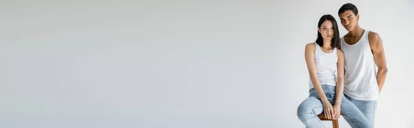 Morena asiático mulher no branco tanque superior e jeans sentado no banquinho perto africano americano cara isolado no cinza, banner — Fotografia de Stock