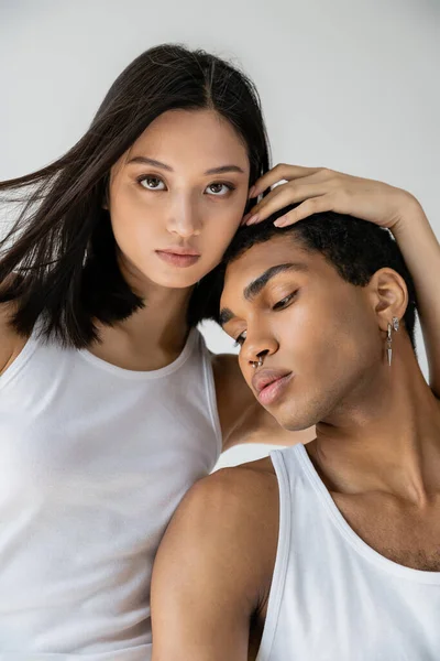 Brunette asian woman in white tank top looking at camera near african american guy with piercing isolated on grey — Stock Photo