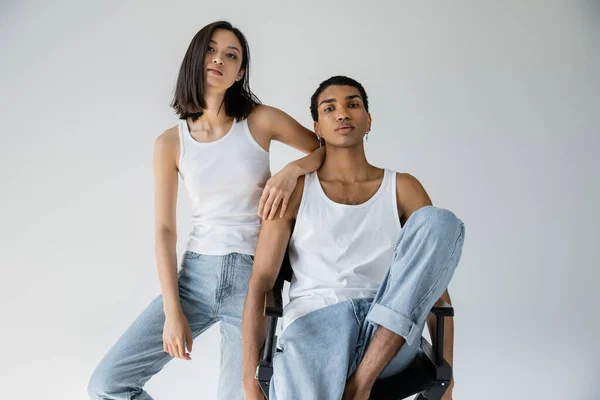 Slender asian woman in blue jeans and white tank top leaning on african american man sitting on chair isolated on grey — Stock Photo