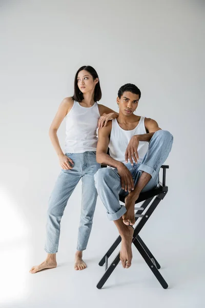 Pleine longueur de pieds nus asiatique femme en débardeur et jeans regarder loin près afro-américain gars assis sur chaise sur fond gris — Photo de stock