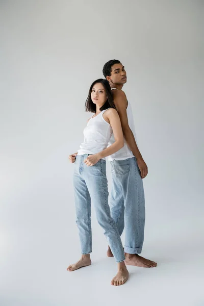 Piena lunghezza della coppia interrazziale a piedi nudi in canotte bianche e jeans in posa back to back su sfondo grigio — Foto stock
