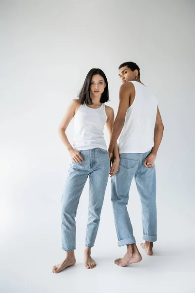 Longitud completa de descalzo asiático mujer posando con el pulgar en bolsillo de azul jeans cerca africano americano chico en gris fondo - foto de stock