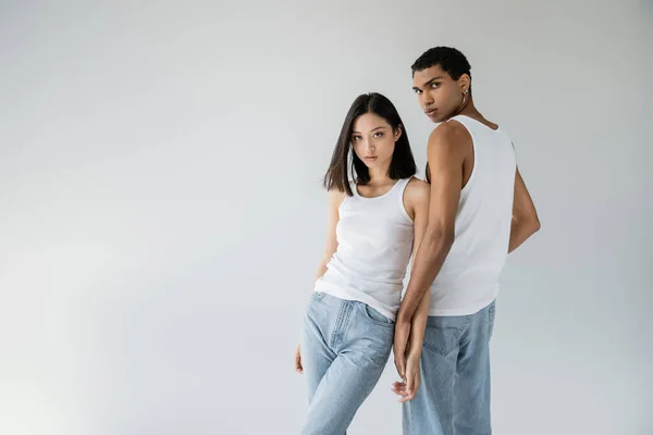 Брюнетка азиатка и африканский американец в синих джинсах и белых майках, смотрящий на камеру, изолированную на сером — стоковое фото