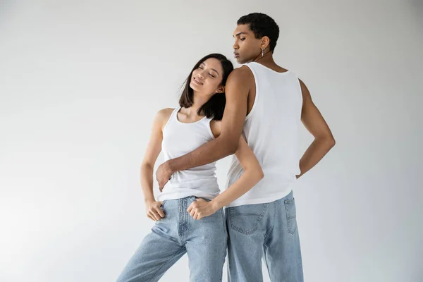 African american man in white tank top and jeans embracing pleased asian woman smiling with closed eyes isolated on grey — Stock Photo