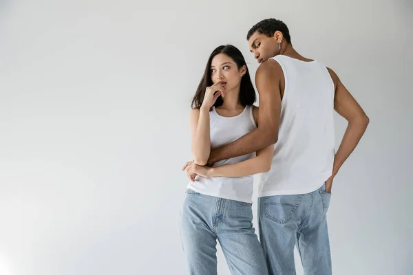 African american man in white tank top and jeans embracing dreamy asian woman looking away isolated on grey — Fotografia de Stock