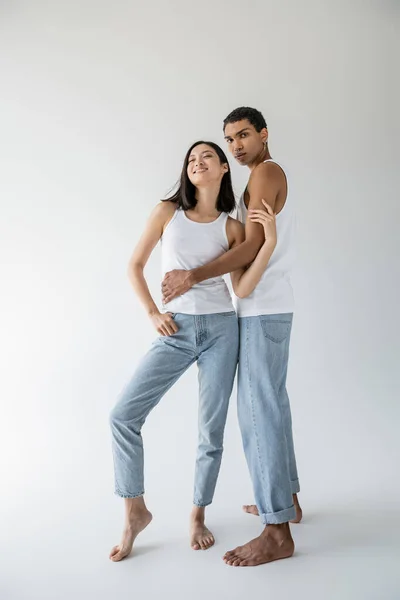 Full length of african american man in tank top and jeans embracing cheerful asian model on grey background — Stock Photo
