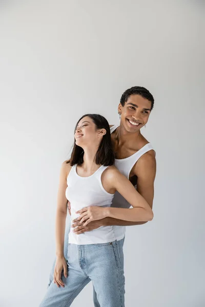 Happy african american guy hugging asian woman smiling with closed eyes isolated on grey — Stock Photo