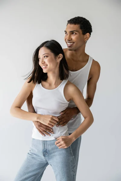 Young african american man embracing carefree asian model in jeans and white tank top isolated on grey — Stock Photo