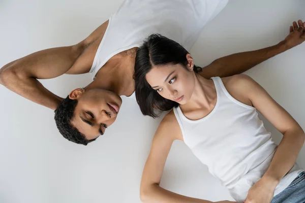 Top view of brunette asian woman and young african american man in white tank tops lying on grey background — Stock Photo