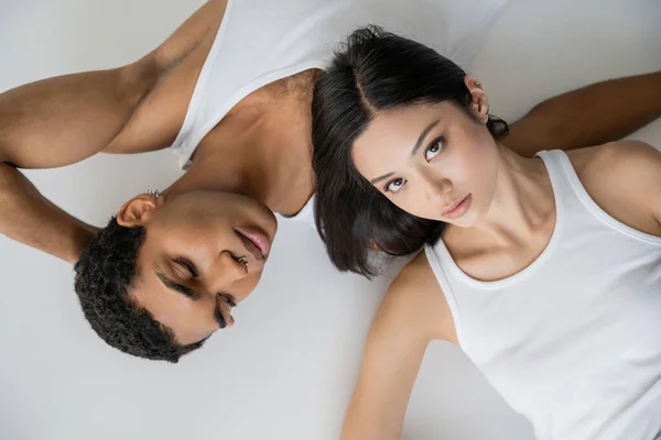 Vue du dessus de la femme asiatique brune en débardeur blanc regardant la caméra près de l'homme afro-américain couché sur fond gris — Photo de stock