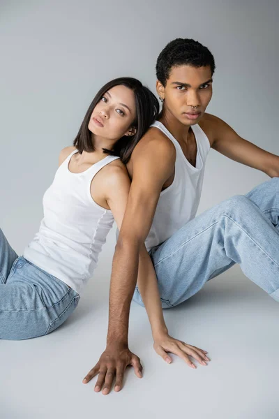 Elegante pareja multiétnica en pantalones vaqueros azules y camisetas sin mangas sentado espalda con espalda sobre fondo gris - foto de stock