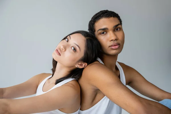 Brunette asian woman and stylish african american man with silver piercing sitting back to back isolated on grey — Stock Photo