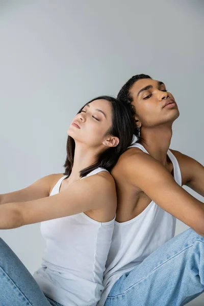 Giovane e trendy coppia interrazziale in canotte bianche seduta schiena a schiena su sfondo grigio — Foto stock
