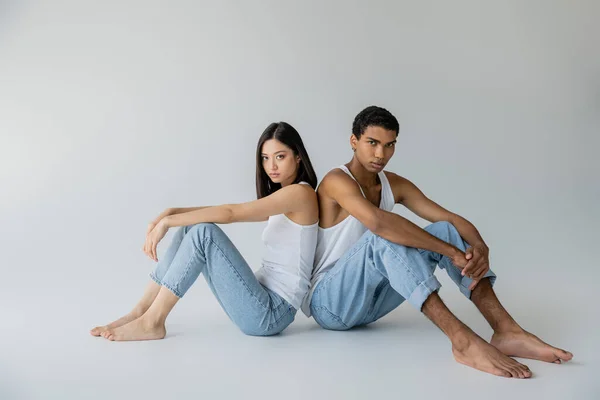 Piena lunghezza di piedi nudi ed elegante coppia interrazziale in jeans e canottiere seduta schiena a schiena su sfondo grigio — Foto stock