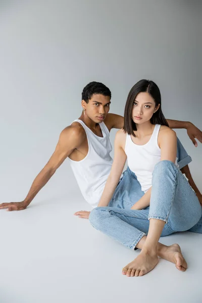 Модная межрасовая пара в джинсах и топах сидит и смотрит на камеру на сером фоне — стоковое фото